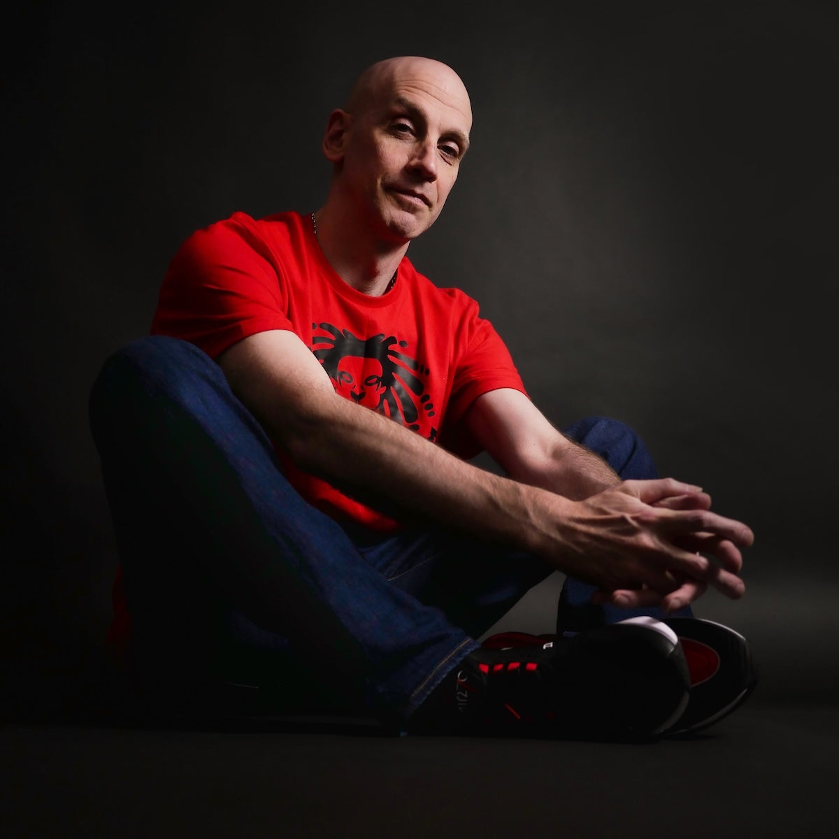 Gav Pauze wearing a red Pauzewear t-shirt with a black graphic design, sitting against a dark background