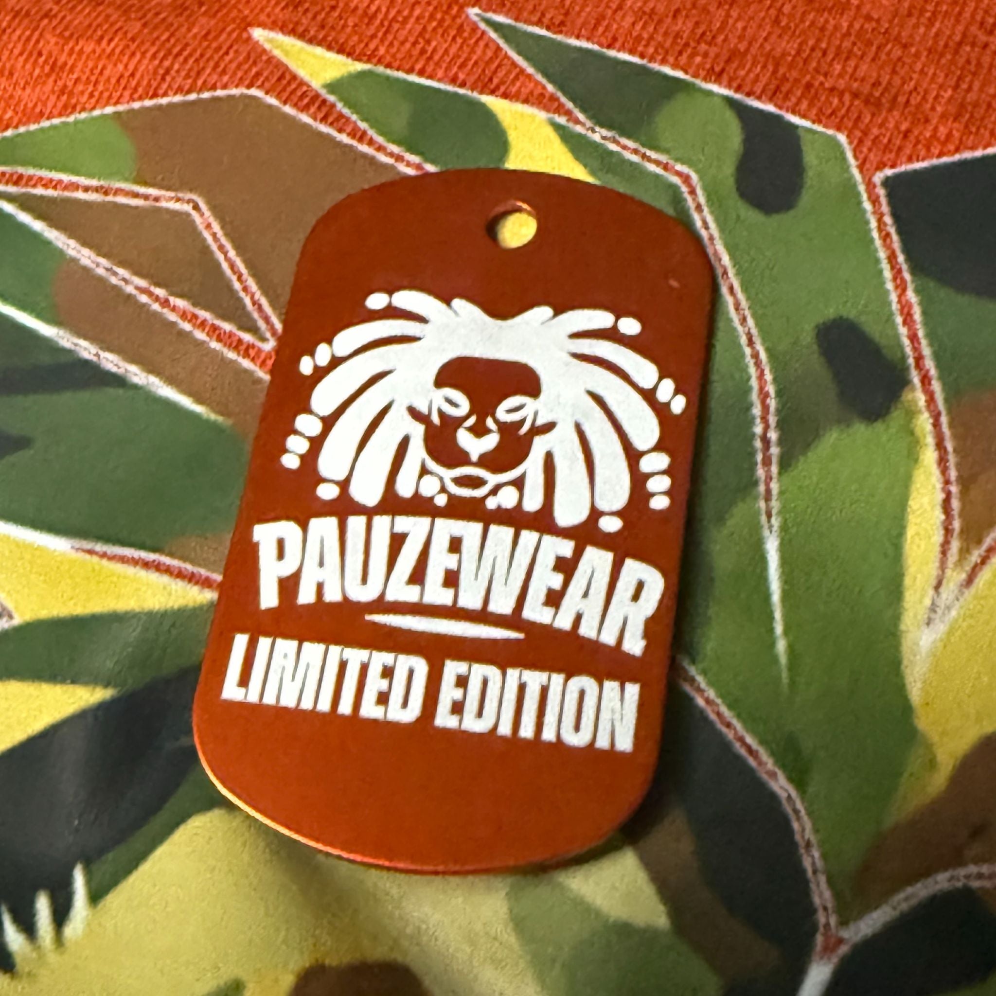 Orange tag with white lion logo and 'PAUZEWEAR LIMITED EDITION' text on a camouflage background