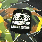 Black dog tag with white lion design and 'PAUZEWEAR LIMITED EDITION' text on a camouflage background