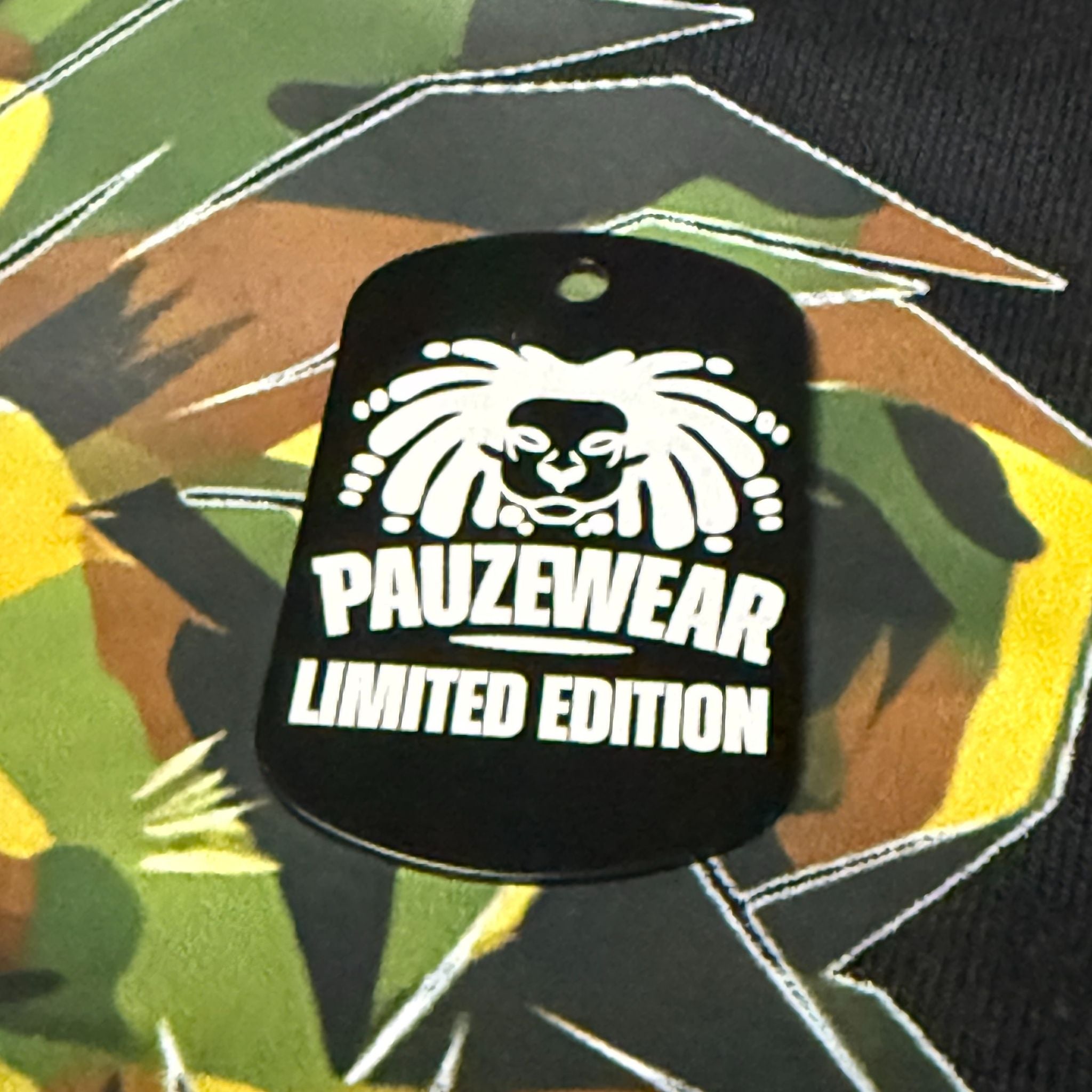 Black dog tag with white lion design and 'PAUZEWEAR LIMITED EDITION' text on a camouflage background